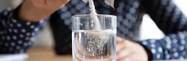 Close up young indian woman pouring soluble anti-influenza powder in glass of water. Sick millennial girl student feeling unwell, relieving grippe fever flu symptoms, high temperature, headache.