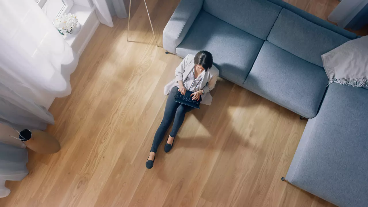 Young Woman is Sitting on a Floor, Working or Studying on a Laptop. Cozy Living Room with Modern Interior, Grey Sofa and Wooden Flooring. Top View Camera Shot.