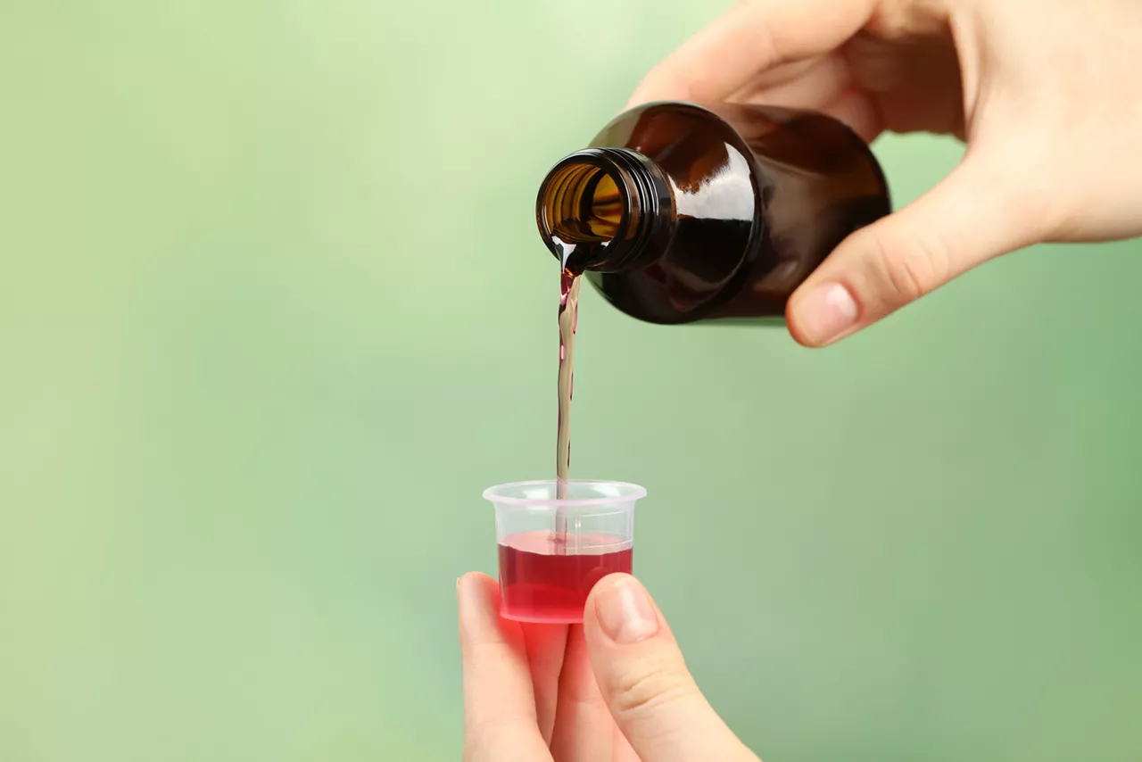 Person pouring cough syrup into measuring cup on light green background